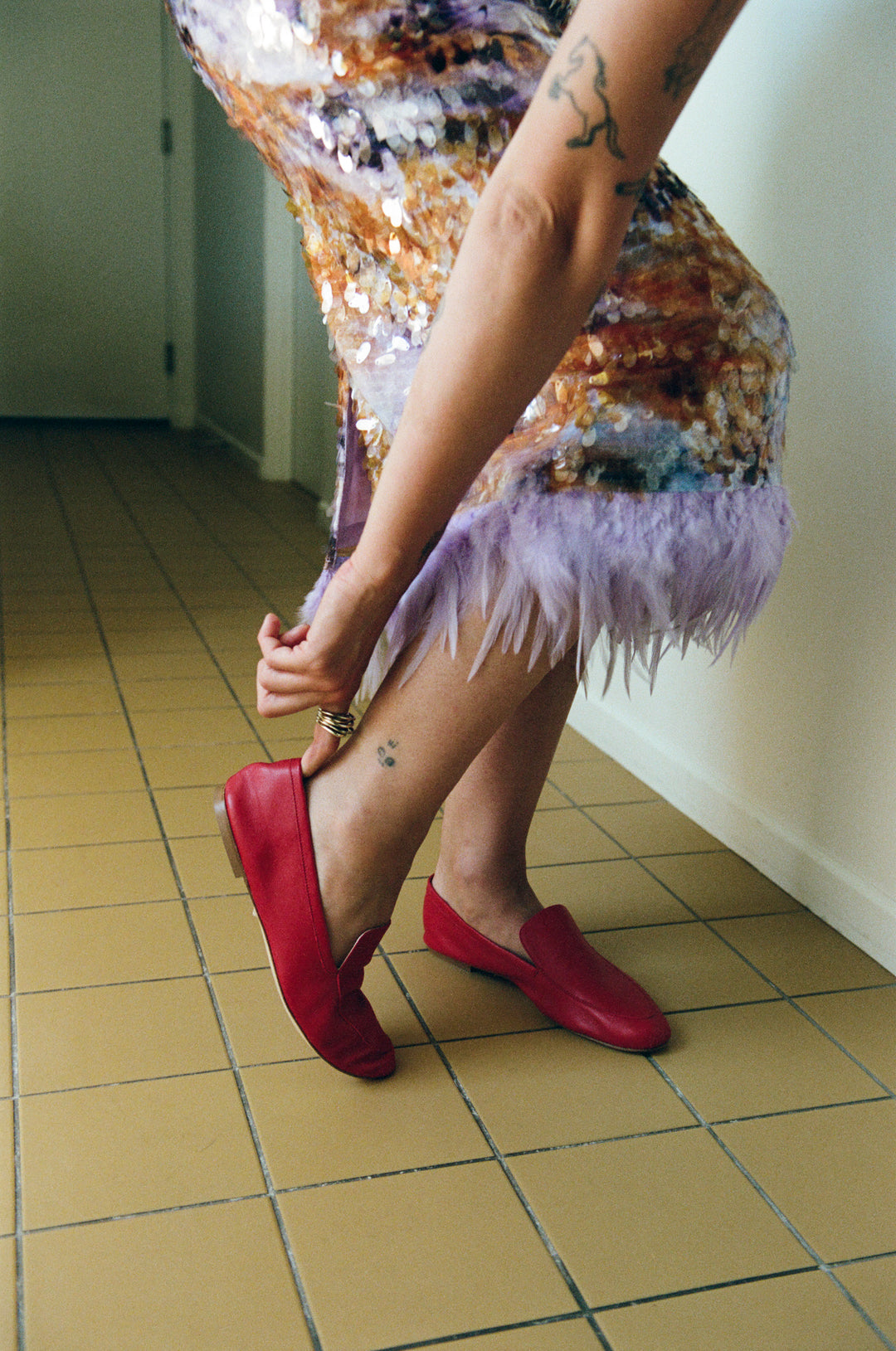 A person wearing a colorful sequined dress with a lavender feather trim and bright red slip-on shoes, standing on a beige tiled floor in an indoor hallway.