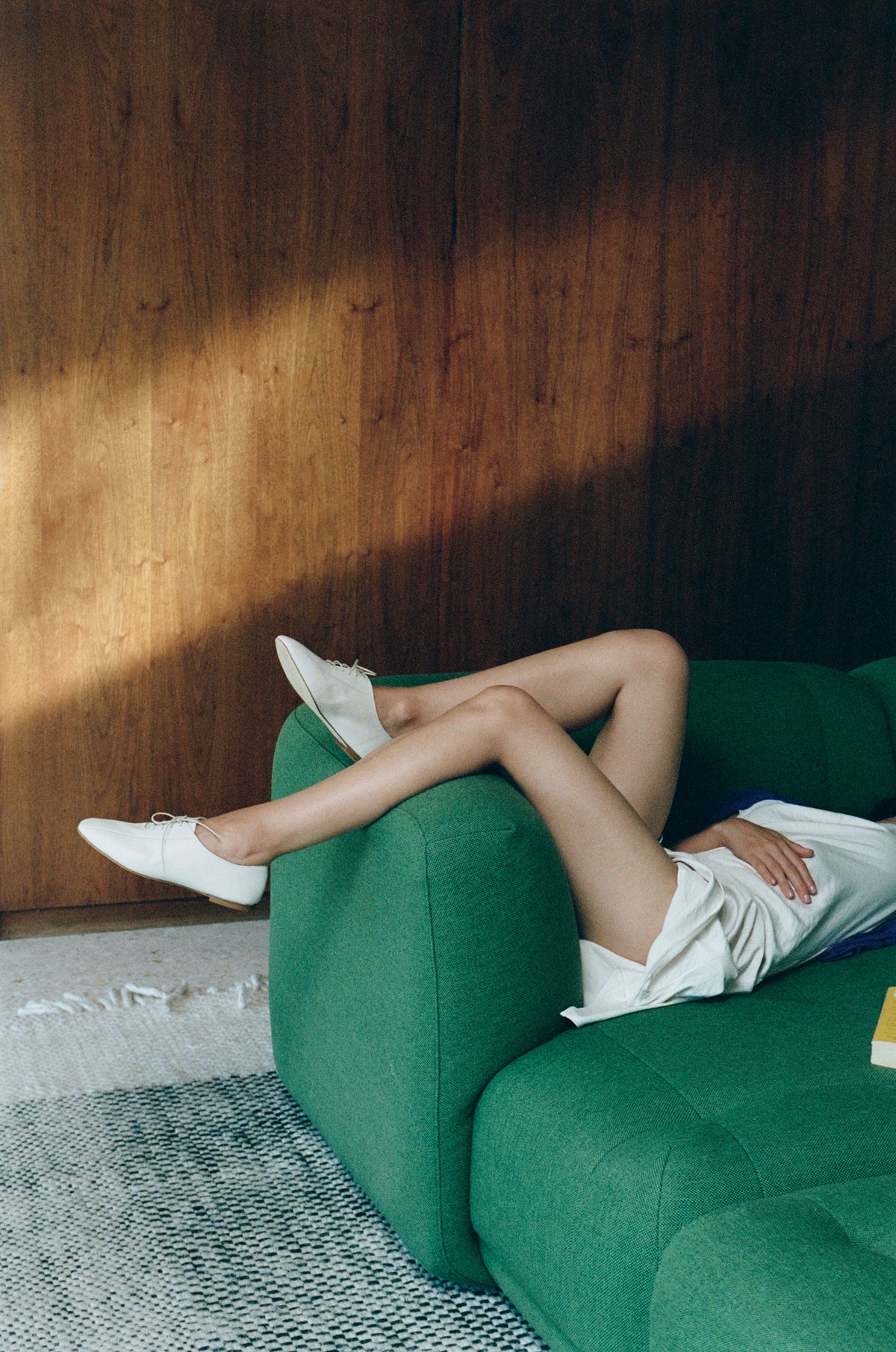 A person reclining on a green sofa with one leg raised, wearing white pointed-toe loafers and a white skirt, set against a dark wood-paneled wall with dramatic sunlight and shadows.